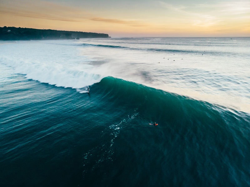 A surfer rides a large ocean wave at dawn with a coastal cliff and other surfers visible.