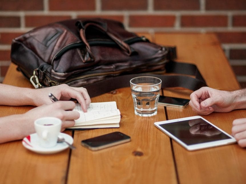 Two people sit at a wooden table with a notebook, smartphone, tablet, coffee cup, and glass.