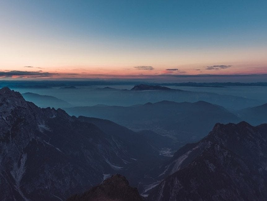 Layered mountain ranges at twilight with a pink and blue sky above misty valleys.